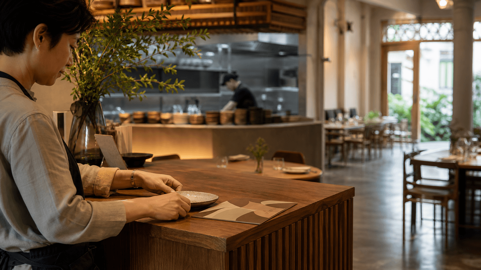 A Singapore restaurant host preparing the dining room before evening service, with the kitchen pass and menu station in the background.